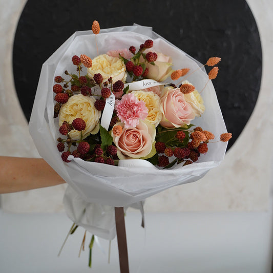 A hand-held bouquet of flowers wrapped in white tissue paper, featuring pale pink and cream roses, small red berries, and dried orange stems, set against a modern wall with a large black circular design.