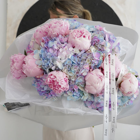 Luxury bouquet of pink peonies and blue hydrangeas wrapped in white paper.