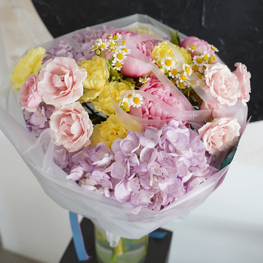 A wrapped bouquet of pink roses, light purple hydrangeas, yellow carnations, and small white daisy-like flowers, photographed from above on a black marble-like surface.