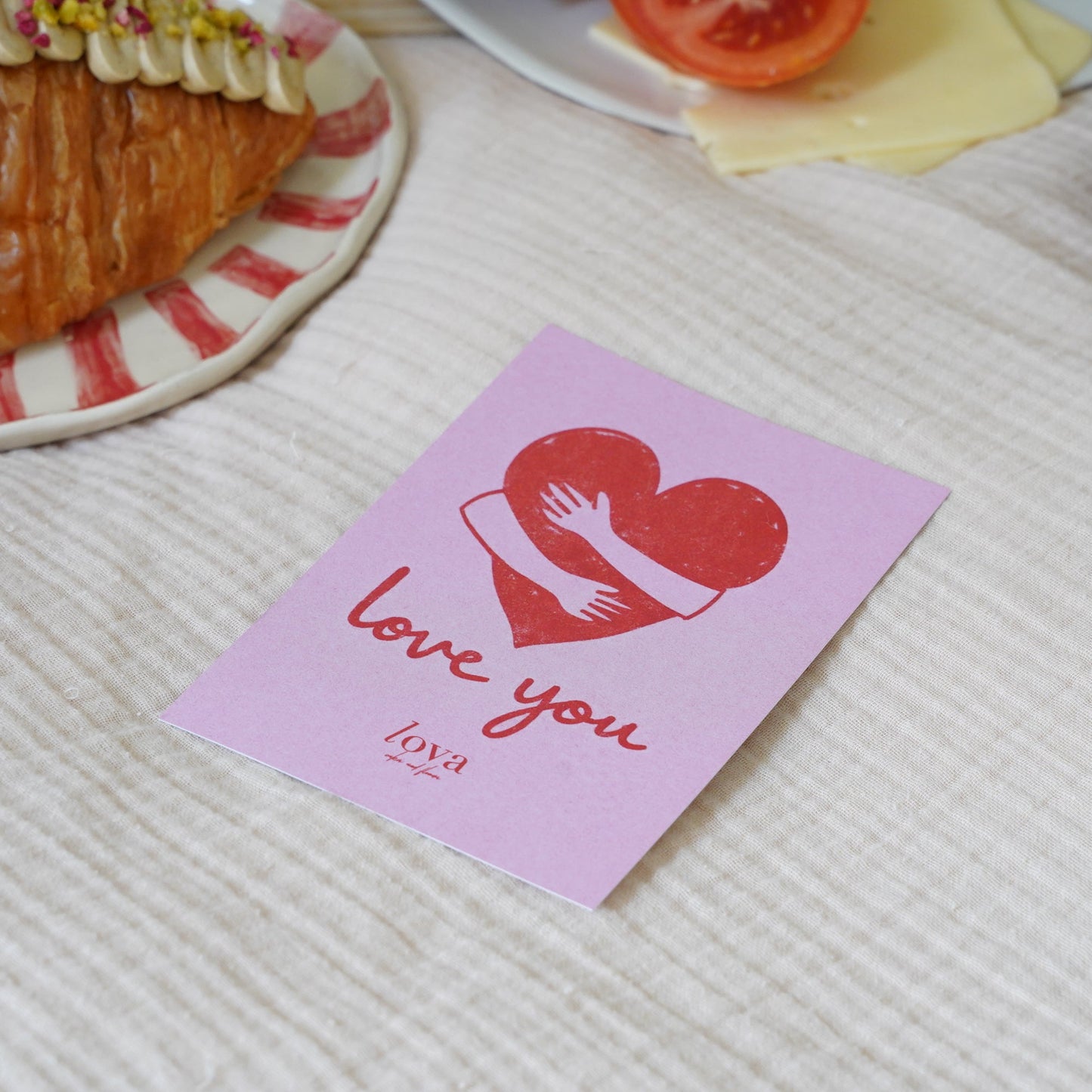 Pink card with heart and hand design on a textured surface with a croissant in the background